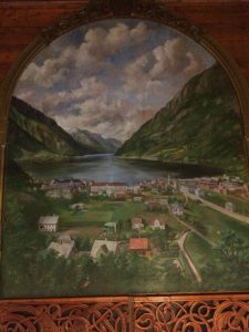 Biletet visar maleriet som heng over scena i Odda ungdomshall. Maleriet er av Odda i gamle dagar med fjorden i bakgrunnen.